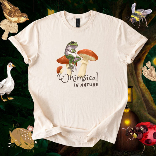 A natural color t-shirt with a print of a frog sitting on a mushroom with its hands crossed, with a caption "whimsical in nature". The shirt is flat laid on a background of a magical forest with whimsical creatures surrounding it.